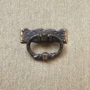 May include: A bronze-toned drawer pull with a decorative ring handle. The rectangular backplate has a hammered texture and is mounted on a neutral-colored woven surface. The ring handle has a twisted design.