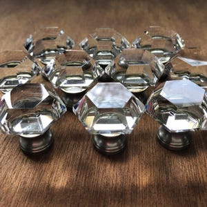 May include: A collection of ten clear, faceted crystal cabinet knobs with a silver-toned base. The knobs are arranged on a wooden surface, showcasing their geometric design and reflective qualities. These decorative knobs are suitable for furniture.