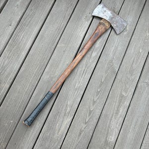 May include: A double-bit axe featuring a weathered, metallic silver head with visible signs of wear and a long, smooth, brown wooden handle. The bottom portion of the handle is wrapped in black tape, providing a secure grip for the user.