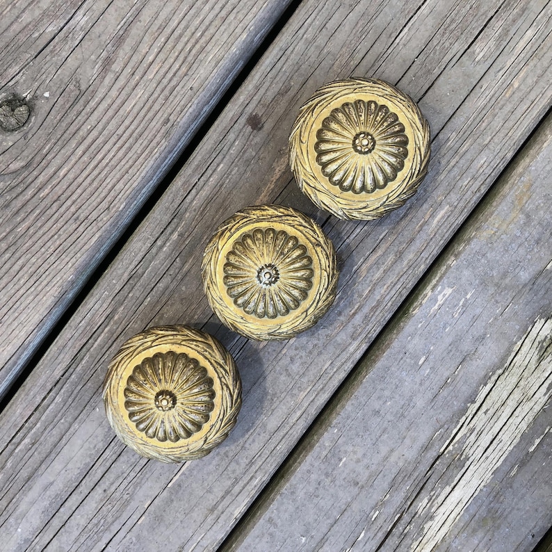 May include: Three antique brass cabinet knobs with a floral design. Each knob features a central flower surrounded by detailed, textured patterns. The knobs are circular and have a warm, aged appearance, suggesting vintage craftsmanship.