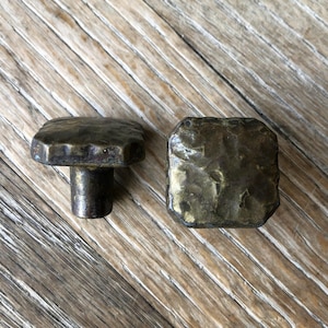 May include: Two hammered metal cabinet knobs with a distressed finish. The knobs are square shaped and have a slightly irregular surface.