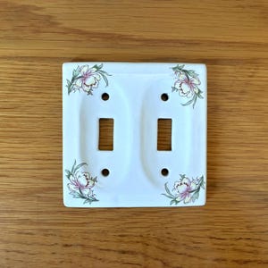 May include: A white rectangular double light switch wall plate featuring two vertical toggle openings and four screw holes. The plate is decorated with delicate floral illustrations in shades of pink, yellow, and green positioned at each corner.