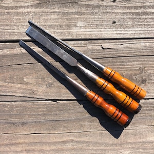 May include: Three vintage wood chisels with wooden handles and metal blades are arranged on a weathered wooden surface. The handles are a warm, reddish-brown color with black stripes, and the blades are silver.