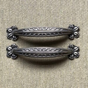 May include: Two antique silver-toned cabinet pulls. Each pull has a central oval handle with a leaf design and is flanked by decorative floral accents. The pulls are displayed on a textured, neutral-toned surface.