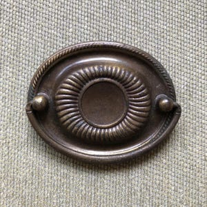 May include: Oval-shaped antique bronze drawer pull with a rope-like border. The center features a circular design with concentric ridges. Small, round handles are on each side of the pull. This hardware is suitable for cabinets and drawers.