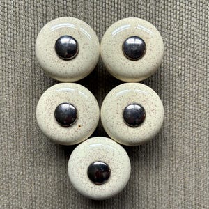 May include: Five round cabinet knobs with a cream-colored ceramic base featuring small brown speckles. Each knob has a smooth, glossy finish and a polished silver-tone metal center cap that is circular and slightly domed in shape.