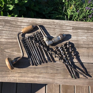 May include: A collection of vintage woodworking tools, including a hand drill and various drill bits, arranged on a weathered wooden surface. The hand drill features a wooden handle and a metal frame. The drill bits are of different sizes and shapes.