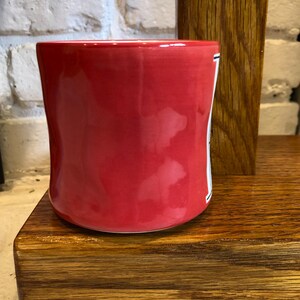May include: A vibrant red ceramic mug with a slightly curved shape. The mug has a glossy finish and a white stripe with black detailing on the side. It sits on a wooden surface, with a brick wall in the background.