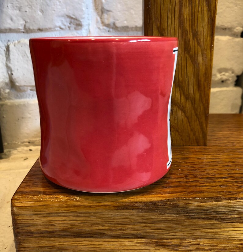 May include: A vibrant red ceramic mug with a slightly curved shape. The mug has a glossy finish and a white stripe with black detailing on the side. It sits on a wooden surface, with a brick wall in the background.