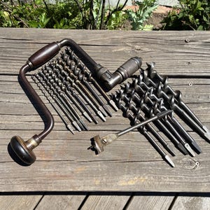May include: A vintage metal hand brace drill with a dark wooden handle and knob, accompanied by a collection of various metal auger drill bits and a single flat spade bit, all arranged together on a weathered wooden surface.