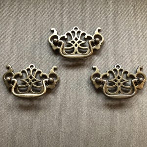 May include: Three antique brass drawer pulls with ornate, symmetrical designs. Each pull features a curved handle and decorative detailing, perfect for adding a vintage touch to furniture. The pulls are set against a neutral background.