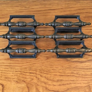 May include: Six antique brass cabinet pulls with a rectangular plate and a decorative, fluted,  handle. The pulls are arranged in two rows of three.