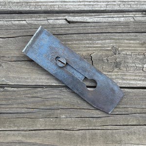 May include: A rectangular metal plane iron with a dark, weathered patina and visible surface oxidation. The tool features a central longitudinal slot with a flat-head screw mechanism and a rounded cutout at the base for adjustment purposes.