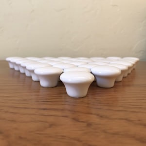 May include: A group of 25 white ceramic knobs arranged in a triangular pattern on a wooden surface.