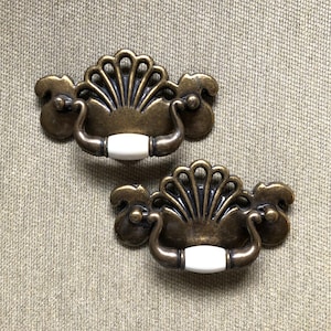May include: Two antique brass cabinet pulls with a decorative fan design and white ceramic knobs.