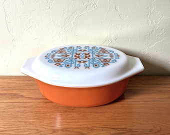 May include: A round orange casserole dish with a white lid featuring a symmetrical geometric pattern in blue and brown. The dish has integrated side handles and a smooth finish, providing a functional vessel for food preparation and serving.