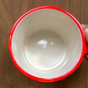 May include: A red-rimmed mug with a cream-colored interior. The mug has a bright red rim and handle, and the interior is a neutral cream color. The mug is empty and appears to be made of ceramic or a similar material.