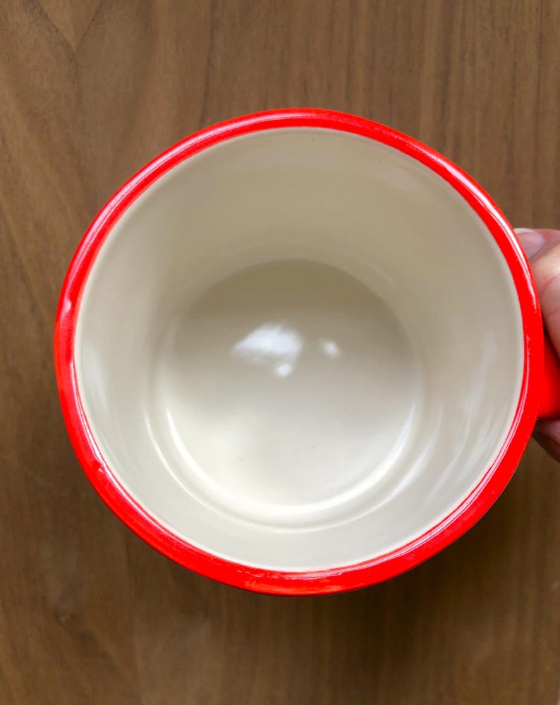 May include: A red-rimmed mug with a cream-colored interior. The mug has a bright red rim and handle, and the interior is a neutral cream color. The mug is empty and appears to be made of ceramic or a similar material.