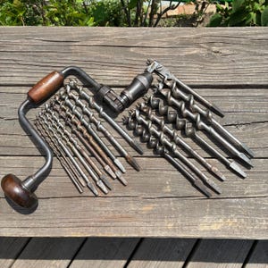 May include: A metal hand brace drill with a dark brown wooden handle and knob, accompanied by a collection of twelve various sized metal auger drill bits arranged in a row, featuring spiral flutes and pointed tips for manual woodworking tasks.