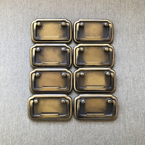 May include: Eight antique brass drawer pulls arranged in a grid. Each pull has a rectangular face with a curved handle and two small, round rivets. The pulls have a warm, aged finish, suggesting vintage or retro style.