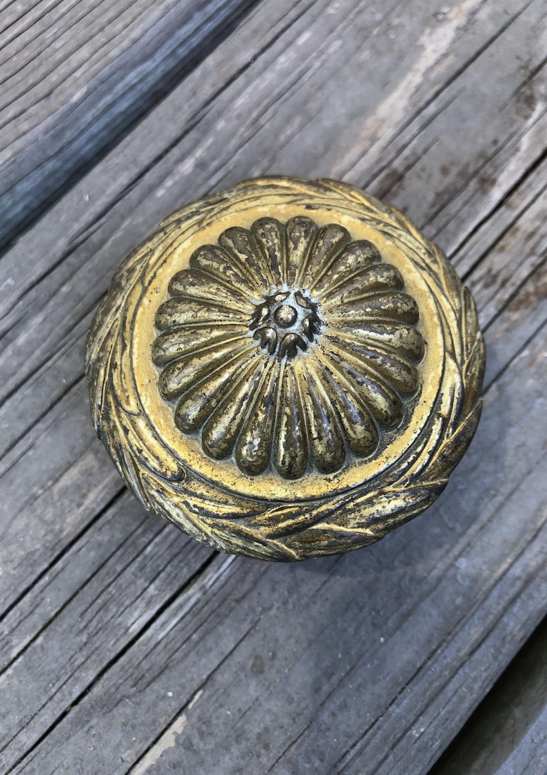 May include: A round, antique brass-colored decorative object with a floral design in the center and a laurel wreath border. The object is set against a weathered wooden surface.