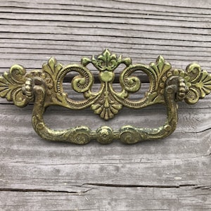 May include: Ornate gold-toned metal drawer pull with a decorative scroll design. The pull has a loop handle.