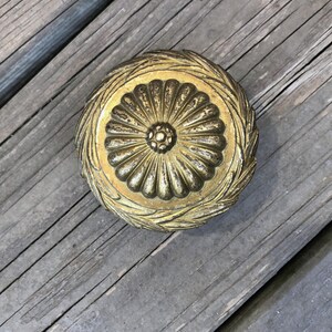 Op de afbeelding: Een rond, antiek ogend decoratief object met een gouden afwerking. Het midden heeft een bloemmotief, omringd door een lauwerkrans. Het object ligt op een verweerd houten oppervlak.