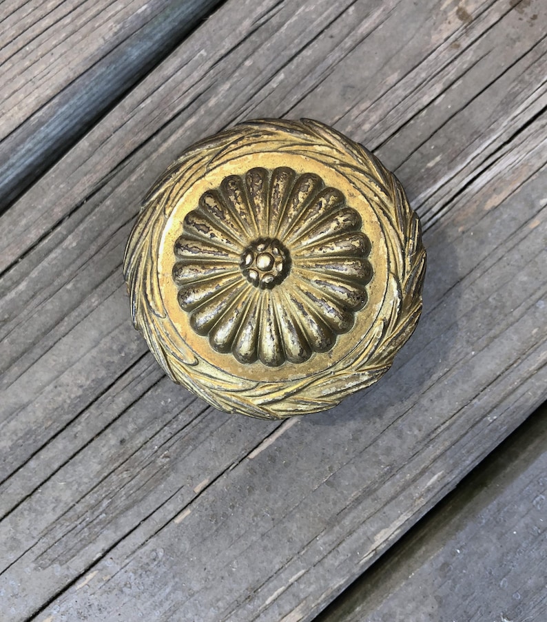 Op de afbeelding: Een rond, antiek ogend decoratief object met een gouden afwerking. Het midden heeft een bloemmotief, omringd door een lauwerkrans. Het object ligt op een verweerd houten oppervlak.