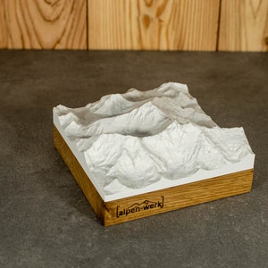 Könnte beinhalten: Ein weißes 3D-gedrucktes Modell einer Bergkette auf einem Holzsockel. Der Sockel ist aus Eichenholz und trägt die Gravur "alpen-werk".