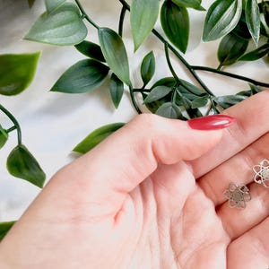 Może przedstawiać: Para srebrnych kolczyków z geometrycznym wzorem atomu. Kolczyki są pokazane na dłoni z czerwonym lakierem do paznokci.