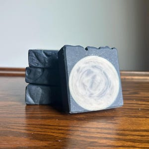 May include: A stack of four rectangular bars of dark charcoal-colored soap. The front-most bar features a prominent, circular, light-colored swirl pattern in the center that resembles a full moon against a dark, textured background.