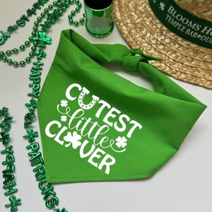 Peut inclure: Un bandana vert vif pour chien avec l'inscription blanche "Cutest Little Clover". Le bandana est noué en haut. L'image comprend également des perles vertes, un chapeau et un verre à liqueur vert, tous sur le thème de la Saint-Patrick.
