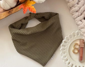 May include: Olive green bandana with a textured, quilted pattern. The bandana is knotted at the top and has a small white tag on the bottom right corner. The bandana is triangular in shape and is likely for a pet.