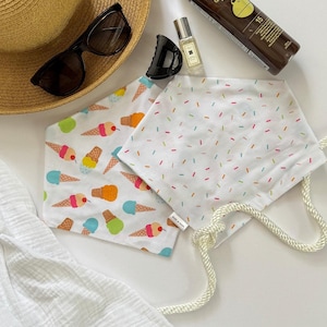 May include: Two white face masks with colorful patterns. One features ice cream cones, the other has sprinkles. A straw hat, sunglasses, and other accessories are arranged around the masks.