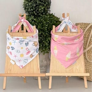 May include: Two triangular fabric bibs displayed on wooden easels. The left bib is white with a colorful summer-themed print of suns, starfish, and beach bags. The right bib is solid pink with a repeating pattern of white ice cream cones.