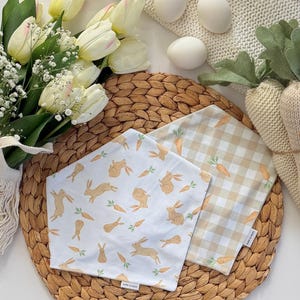 May include: Two baby bibs with Easter-themed patterns rest on a woven mat. One bib features a white background with tan bunnies and carrots. The other has a tan and white gingham pattern with carrots. A bouquet of white tulips and eggs are nearby.