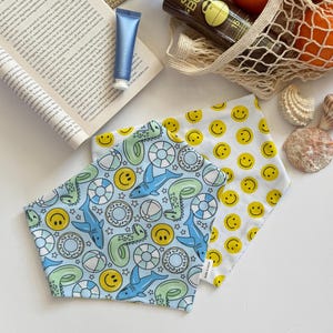 May include: Two blue and white bandana bibs with a summer theme. One bib has a pattern of sharks, donuts, and beach balls. The other bib has a pattern of yellow smiley faces.