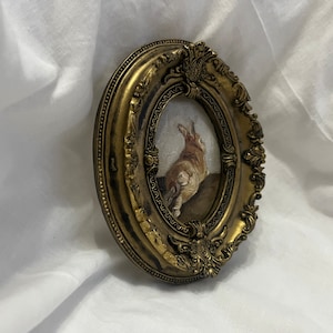 Puede incluir: Un marco ovalado dorado con detalles ornamentados rodea una pintura de un conejo marr&oacute;n mirando hacia arriba. El conejo est&aacute; de pie sobre una superficie marr&oacute;n.