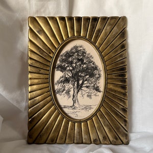 Peut inclure: Un cadre photo ovale doré avec un motif en forme de soleil. Le cadre contient un dessin en noir et blanc d'un arbre.