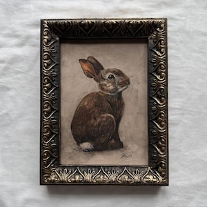 Peut inclure: Une peinture encadrée représentant un lapin brun avec une queue blanche, vu de profil sur un fond neutre. L'œuvre est insérée dans un cadre métallique sombre et orné, doté d'un motif en relief répété sur les bords et d'une signature en bas.