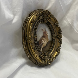 Puede incluir: Un marco ovalado dorado con detalles ornamentados rodea una pintura de un conejo marr&oacute;n mirando hacia arriba. El conejo est&aacute; de pie sobre una superficie marr&oacute;n.