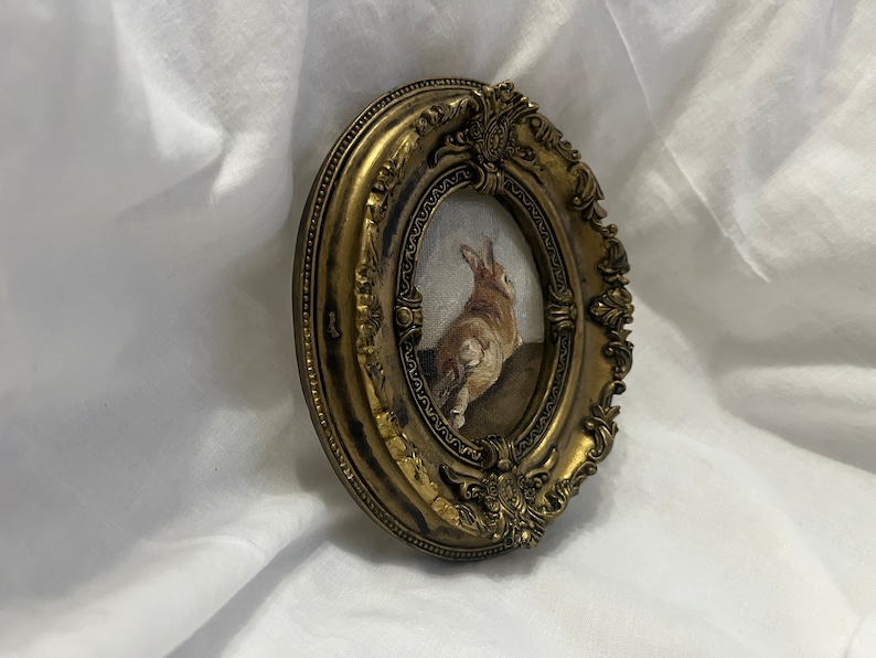 Puede incluir: Un marco ovalado dorado con detalles ornamentados rodea una pintura de un conejo marr&oacute;n mirando hacia arriba. El conejo est&aacute; de pie sobre una superficie marr&oacute;n.