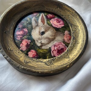 May include: A white rabbit with pink eyes is painted in a gold frame with pink roses and green leaves. The frame is round and has a textured surface.