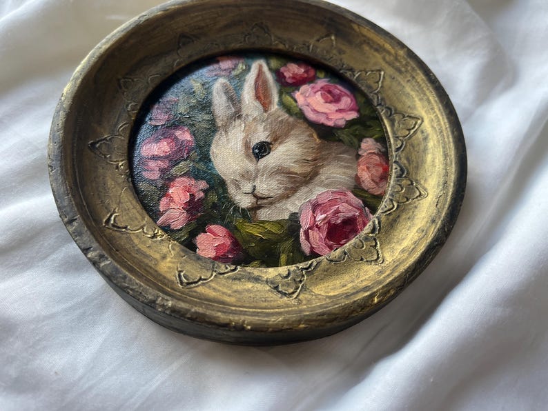 May include: A white rabbit with pink eyes is painted in a gold frame with pink roses and green leaves. The frame is round and has a textured surface.