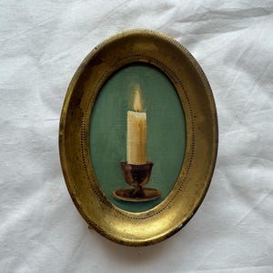 May include: Oval-shaped painting of a lit candle in a dark brown holder, set against a teal background. The painting is framed in a gold-colored, ornate oval frame. The candle has a yellow flame.