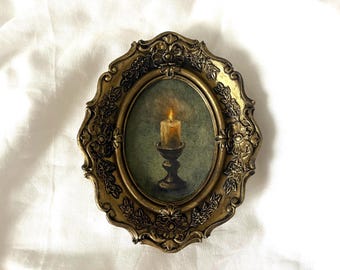 May include: Oval-shaped framed artwork featuring a lit candle on a pedestal. The painting has a green background and is set within an ornate, gold-colored frame with floral detailing. The candle flame glows brightly.