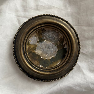 以下が含まれることがあります： 小さな丸いアンティーク真鍮製のフォトフレーム、ビーズの縁取り付き。 フレームには、グレー、グリーン、ブラウンのくすんだ色合いの水彩画が収められています。 フレームは白い布の背景に置かれています。