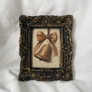 Pode incluir: Uma pintura em aquarela de um sino com um laço marrom, emoldurada em uma moldura dourada ornamentada.