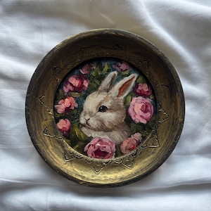 May include: A round, gold-colored frame with a painted image of a white rabbit surrounded by pink roses. The rabbit is looking to the left and has a pink nose.