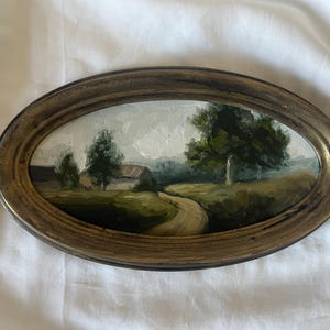 May include: A framed oil painting depicting a rural landscape with a winding dirt road leading to a small white farmhouse. The painting features a large green tree in the foreground and a cloudy sky.